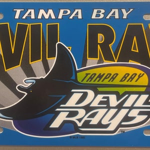 May include: A blue and yellow Tampa Bay Devil Rays license plate with a black stingray logo. The text "Tampa Bay Devil Rays" is printed in black with a yellow outline.