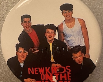 May include: A vintage button featuring a photo of the boy band New Kids on the Block. The five members are wearing casual clothing and are smiling. The band's name is written in red letters below the photo.
