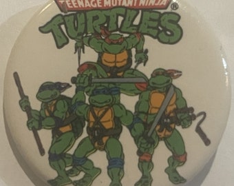 May include: A white round button with a Teenage Mutant Ninja Turtles logo. The logo features the words "Teenage Mutant Ninja Turtles" in green and red lettering. Four green turtles are depicted in the center of the button, each with a different colored bandana. The turtles are holding weapons, including a sai, katanas, and a nunchaku.