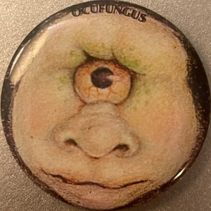 May include: A round button with a cartoon face with one large eye and a large nose. The eye is orange with a black pupil. The nose is flesh-colored. The face is flesh-colored with green spots. The text "OCUFUNGUS" is printed in black at the top of the button.