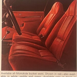 May include: A vintage car interior with red bucket seats. The seats are made of a textured material and have a classic design. A black steering wheel and part of the dashboard are visible. The image is from an advertisement.