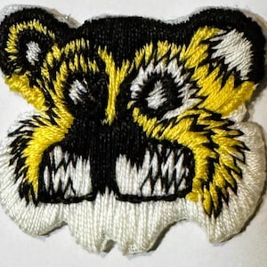 May include: Embroidered patch featuring a stylized, ferocious face with sharp teeth. The design uses black, yellow, and white thread to create a striking, detailed image. The patch has a unique, irregular shape, suggesting it's meant to be sewn onto fabric.