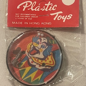May include: A plastic toy with a clown face and a red, yellow, blue, and green design. The toy is in a clear plastic bag with a red and white label that reads "Plastic Toys" and "Made in Hong Kong".