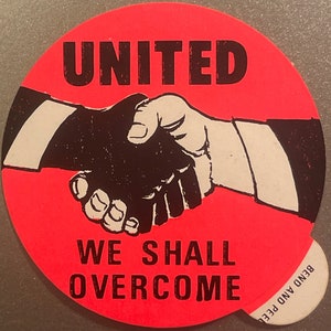 May include: A red circular sticker with the text "UNITED WE SHALL OVERCOME" in black lettering. The sticker features a black and white illustration of two hands shaking.