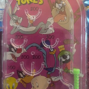 May include: A plastic Looney Tunes pinball game with a pink background. The game features characters from the cartoon, including Bugs Bunny, Daffy Duck, Tweety Bird, Sylvester the Cat, and Tasmanian Devil. The game has a spring-loaded plunger and a variety of targets with point values.