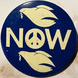 May include: A blue round button with the word "NOW" in white letters and a peace sign in the center. Two white doves with olive branches in their beaks are on either side of the word "NOW".