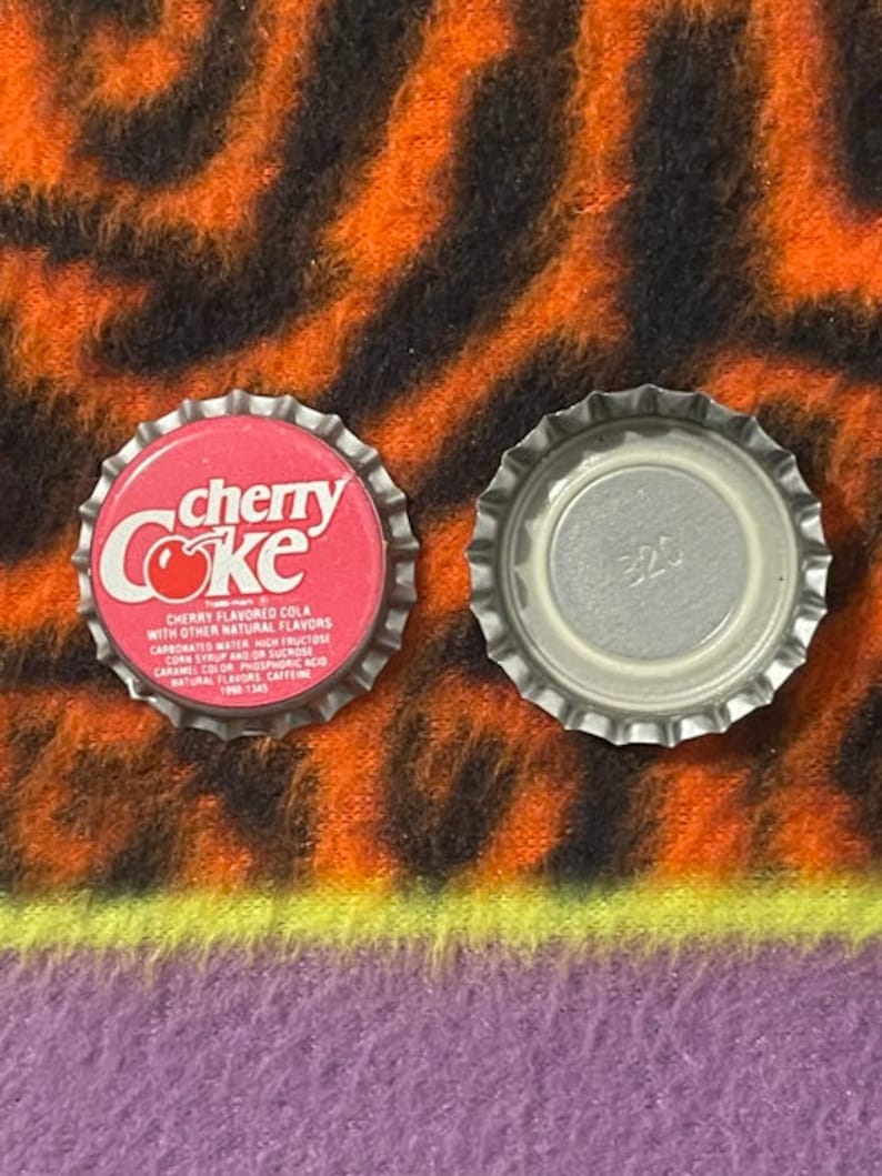 Op de afbeelding: Twee flessendoppen, een rode met de woorden "cherry Coke" en de andere zilveren met het nummer "320" erop.