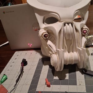 May include: A white, unpainted, full-head helmet with a complex, mechanical design featuring a wide brow, recessed eye sockets, and a prominent, multi-layered jaw structure with circular side details, resting on a grid-patterned cutting mat.