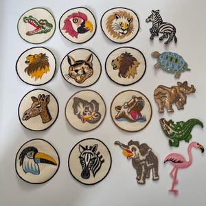 Può includere: Una collezione di toppe ricamate con animali, tra cui una zebra, un leone, una giraffa e un elefante. Le toppe sono circolari con sfondo color crema e bordi neri. Altre toppe includono un tucano, un alligatore e un fenicottero.
