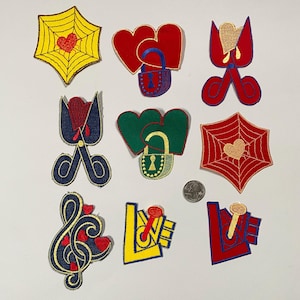 Op de afbeelding: Negen geborduurde strijkpatches met verschillende ontwerpen, waaronder harten, sloten, sleutels, scharen, muzieknoten en een spinnenweb. De patches zijn in rood, geel, blauw, groen en goud.