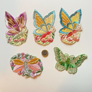 Op de afbeelding: Vijf geborduurde vlinder- en insectenpatches in verschillende kleuren. De patches hebben gedetailleerde vleugels met gouden, blauwe, groene en roze accenten. De patches liggen op een witte ondergrond, met een euromunt als schaal.