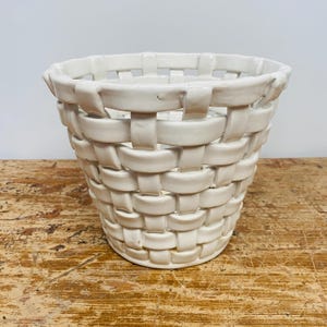 Op de afbeelding: Witte keramische plantenpot met een geweven ontwerp. De pot is gevormd als een mand en heeft een brede, open bovenkant.
