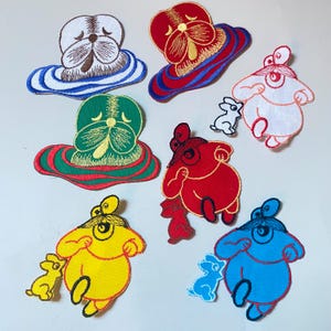 May include: Nine embroidered iron-on patches featuring cartoon characters. The characters are a white, red, green, yellow, and blue creature with a rabbit beside each. The characters are all wearing a similar expression with large eyes and a small mouth.