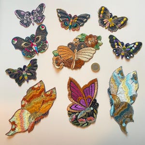 Op de afbeelding: Een verzameling geborduurde vlinderpatches in verschillende maten en kleuren. De patches hebben gedetailleerde stiksels en een verscheidenheid aan kleuren, waaronder zwart, goud, paars en oranje. Sommige patches hebben bloemaccenten.