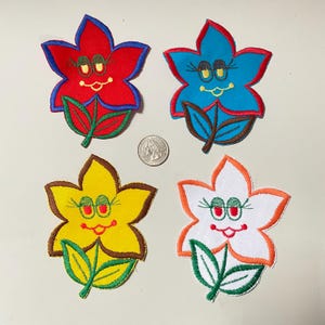 May include: Four colorful flower-shaped embroidered patches. Each patch features a smiling face with eyelashes, a mouth, and a pair of eyes. The flowers are red, blue, yellow, and white, with green leaves and brown outlines.