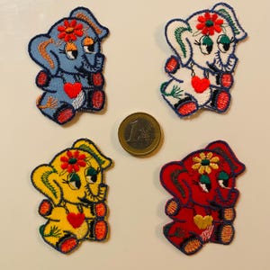 Könnte beinhalten: Vier gestickte Elefantenaufnäher in Blau, Weiß, Gelb und Rot, jeder mit einer Blume auf dem Kopf und einem Herz auf der Brust. Neben den Aufnähern liegt eine Euro-Münze. Die Aufnäher sind farbenfroh und detailliert.