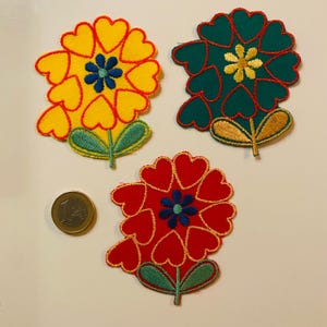 Könnte beinhalten: Drei gestickte Blumenaufnäher in Gelb, Grün und Rot, jeweils mit herzförmigen Blütenblättern und einem zentralen Blumendesign. Neben den Aufnähern liegt eine Euro-Münze. Die Aufnäher haben grüne Stiele und Blätter.