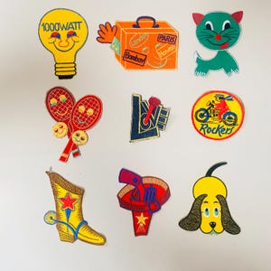 Peut inclure: Un assortiment de patchs brodés avec divers motifs. Comprend une ampoule jaune avec "1000WATT", une valise orange avec des autocollants de voyage, un chat vert, des raquettes de tennis, un microphone, un patch de moto, une botte de cowboy jaune, un chapeau rouge et un chien jaune.
