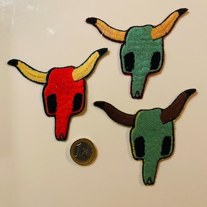 Könnte beinhalten: Drei gestickte Bullenschädel-Patches in Rot, Grün und Grün mit gelben, braunen und schwarzen Hörnern. Neben den Patches liegt eine Euro-Münze. Die Patches haben schwarze Umrisse und liegen auf einer weißen Oberfläche.