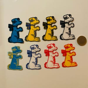 Op de afbeelding: Een verzameling van acht geborduurde patches in de vorm van een hond, in verschillende kleuren, waaronder blauw, geel, wit en rood. Elke hond heeft een contrasterende strik. Een euromunt ligt ernaast.