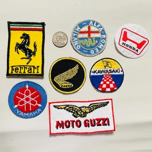Peut inclure: Une collection de patchs brodés représentant différentes marques de motos et de voitures, notamment Ferrari, Honda, Kawasaki, Yamaha, Moto Guzzi et Alfa Romeo. Les patchs sont colorés et présentent des logos et du texte.