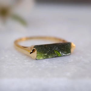 May include: A gold ring featuring a green, rectangular, raw gemstone. The gemstone has a rough, natural texture.