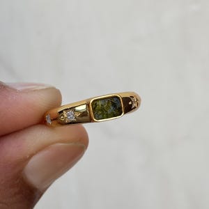 Puede incluir: Un anillo de oro con una piedra rectangular de color verde. El anillo tiene una banda sencilla con pequeñas piedras de acento claras y detalles decorativos. El anillo está sostenido por los dedos de una persona.