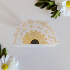 May include: A die-cut sticker featuring a yellow sunflower with a brown center. The sticker has the text "Sweetest of the sunflowers, you're the sun to me, yeah" in a handwritten style around the sunflower.