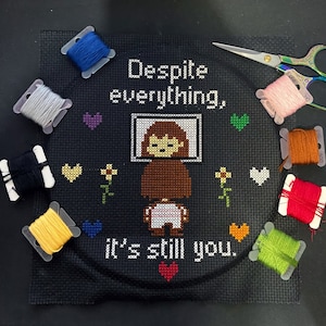 May include: Cross-stitch embroidery on black fabric with the text "Despite everything, it's still you."  The design features a pixelated image of a person with brown hair and a white shirt, surrounded by hearts and flowers.