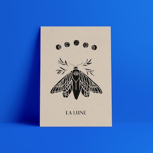 May include: A black and white illustration of a moth with a single eye on its body. The moth is surrounded by five crescent moons. The text "LA LUNE" is printed below the moth.