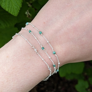 May include: Four delicate silver chain bracelets with small green and white beads. The bracelets are layered on a person's wrist.