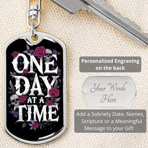 May include: A silver metal keychain with a black and silver dog tag. The dog tag has a black background with a floral design and the text "ONE DAY AT A TIME". The keychain can be personalized with an engraving on the back.