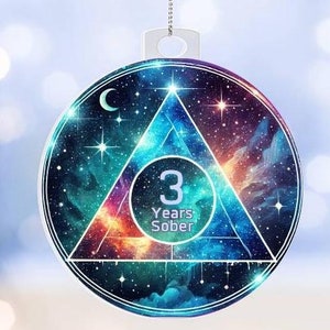 May include: A blue and purple ornament with a triangle design and a galaxy background. The triangle has a white circle in the center with the text "3 Years Sober".