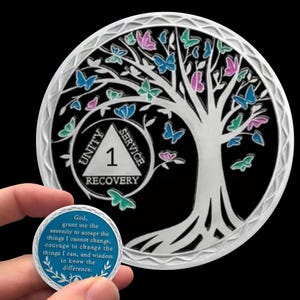 May include: A silver coin with a tree and butterfly design, featuring the words "Unity, Service, Recovery" and the number "1." A smaller coin with a quote about serenity is held in the foreground. The background is black.