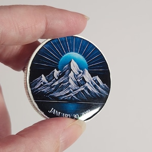May include: A circular, silver-edged item depicting a mountain scene beneath a blue sky and sunburst. The date "JANUARY 30 2019" is inscribed at the base. The artwork is detailed and vibrant.