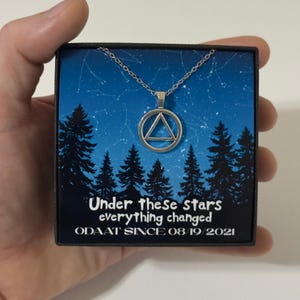 May include: Silver necklace with a circular pendant containing a triangle design. The necklace is presented in a black box with a blue backdrop, pine tree silhouettes, and the text "Under these Stars everything changed ODAAT SINCE 08/19/2021."