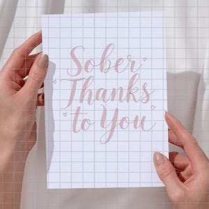 May include: A white card with pink calligraphy that reads "Sober Thanks to You".