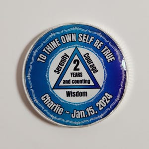 May include: A round, blue and silver commemorative coin with the text "TO THINE OWN SELF BE TRUE". The coin features a triangle with the number "2" and the words "Serenity", "Courage", and "Wisdom". The text "Charlie - Jan. 15. 2024" is also present.