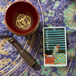 Könnte beinhalten: Eine Tarotkartenlegung mit der Karte Neun der Schwerter nach oben. Die Karte zeigt eine Person, die im Bett liegt und sich den Kopf in die Hände nimmt. Die Karte liegt auf einem lila und grünen gemusterten Stoff mit einer Holz-Singenschale und einem Schlägel.