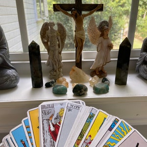 Könnte beinhalten: Eine Tarotkarten-Auslage auf einer weißen Oberfläche mit einer religiösen Szene im Hintergrund. Die Szene zeigt ein Kruzifix mit einer Jesus Christus-Figur, zwei Engeln und mehreren Kristallen. Die Tarotkarten sind in einem Halbkreis ausgelegt, wobei die Rückseiten der Karten und die Vorderseite der Karte Fünf der Stäbe sichtbar sind.