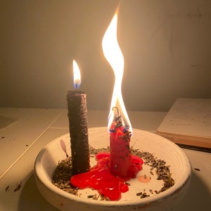 Könnte beinhalten: Zwei brennende Kerzen, eine schwarze und eine rote, stehen in einer weißen Schale mit getrockneten Kräutern. Die rote Kerze schmilzt, das Wachs sammelt sich um ihren Fuß. Flammen steigen von den Kerzen auf und werfen einen warmen Schein.