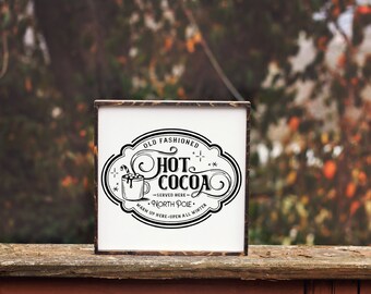 Könnte beinhalten: Ein weißes Schild mit einer schwarz-weißen Illustration einer Tasse heißer Schokolade mit Marshmallows und einem Zuckerstangen. Das Schild lautet "Old Fashioned Hot Cocoa - Served Here - North Pole - Warm Up Here - Open All Winter".