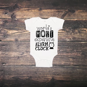 May include: White baby onesie with the text "world's MOST expensive ALARM CLOCK" in black. The onesie has short sleeves and a snap closure. The background is a dark brown wooden surface.
