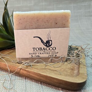May include: A rectangular bar of beige soap with visible flecks, wrapped in a tan paper label featuring a black pipe icon, the text TOBACCO HAND CRAFTED SOAP, and the handwritten weight markings 5.1oz and 145g on the front of the packaging.