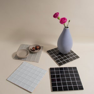 May include: Four square ceramic tiles in different colors: white, gray, black, and dark gray. The tiles are arranged in a grid pattern. A small bowl with two chocolate candies sits on the gray tile. A vase with pink flowers sits on the black tile.