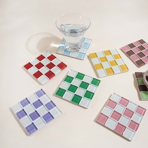 May include: Set of eight square ceramic coasters with a checkered pattern in various colors, including red, yellow, green, purple, pink, and brown. The coasters are arranged on a white surface.