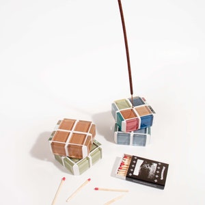 May include: Two stacked square glass incense holders with a brown incense stick standing upright in the top holder. A box of matches with a black and white image of a mountain range is on the white surface. There are five wooden matches with red tips scattered on the surface.