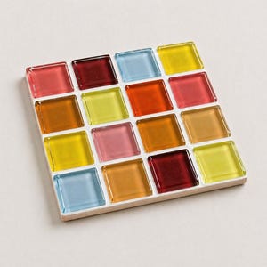 May include: Square coaster with a mosaic tile design. The coaster features sixteen colorful glass tiles in shades of red, orange, yellow, pink, and blue, set on a white base. The tiles are arranged in a grid pattern.