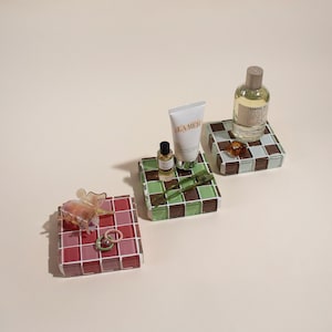 May include: Three small, square, tiled coasters in green, brown, and pink. The coasters are decorated with small, clear glass butterflies, rings, and bottles of perfume. The perfume bottles are labeled "Santal 33" and "La Mer".