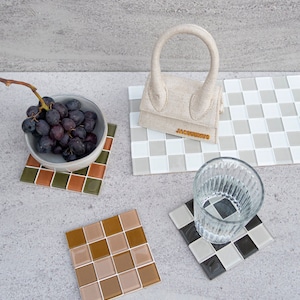 May include: Four ceramic tile coasters in different colors and patterns. One coaster is brown and green, one is brown and beige, one is black and white, and one is gray and white. A small beige handbag with the word "JACQUEMUS" on it is sitting on the gray and white coaster. A glass of water is sitting on the black and white coaster. A bowl of grapes is sitting on the brown and green coaster.